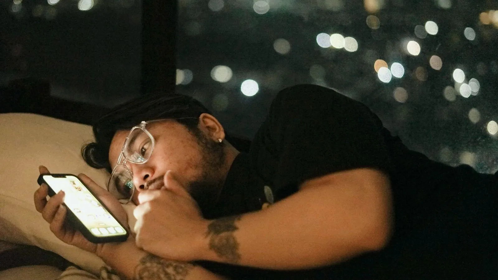 A person in glasses lying on a pillow at night and looking at a phone, with city lights blurred behind.