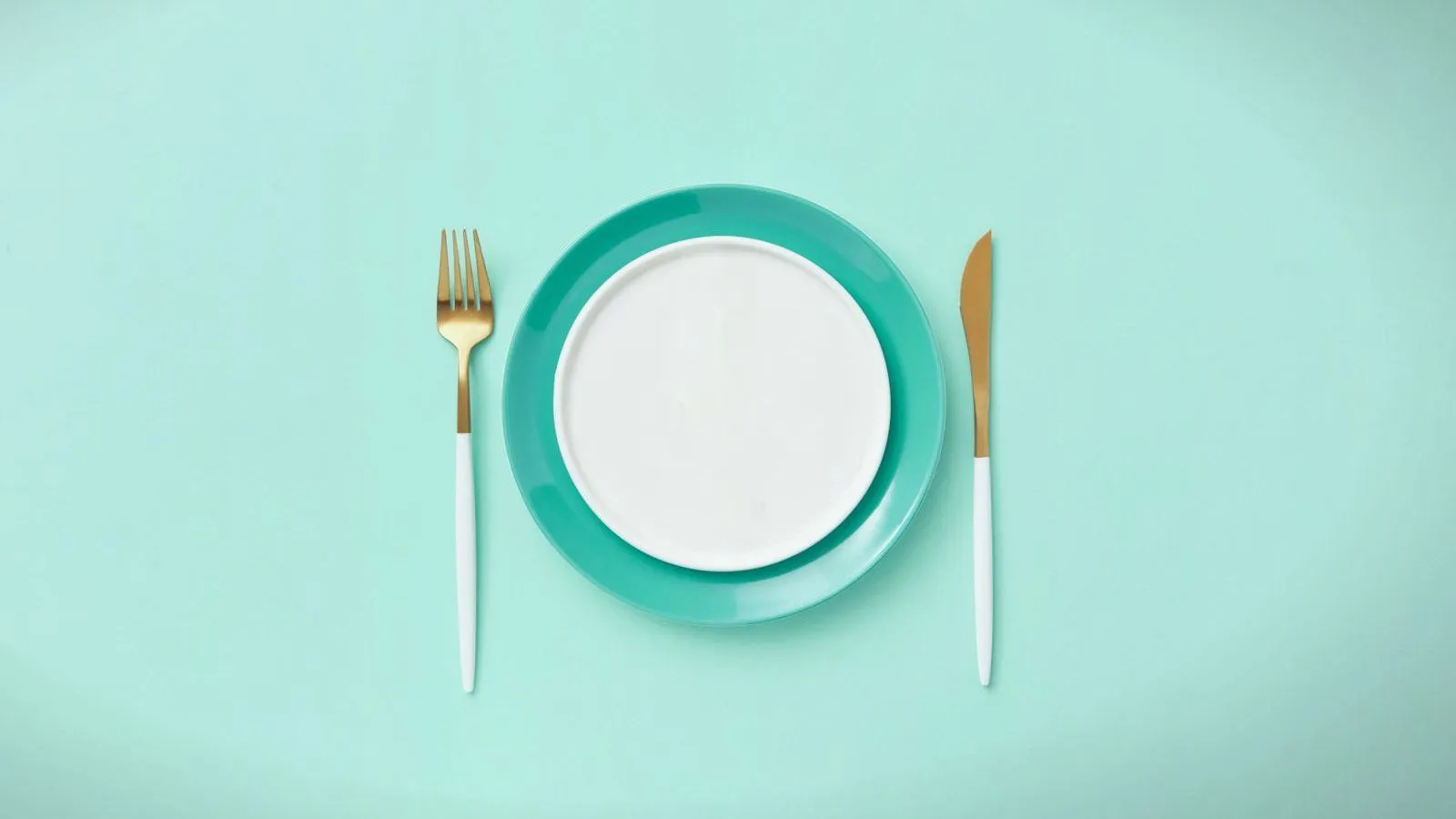 An empty white plate on a teal plate with a fork and knife on a matching teal background.