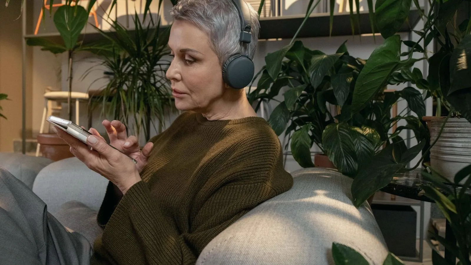 A woman with short gray hair wearing over-ear headphones, using a phone on a sofa