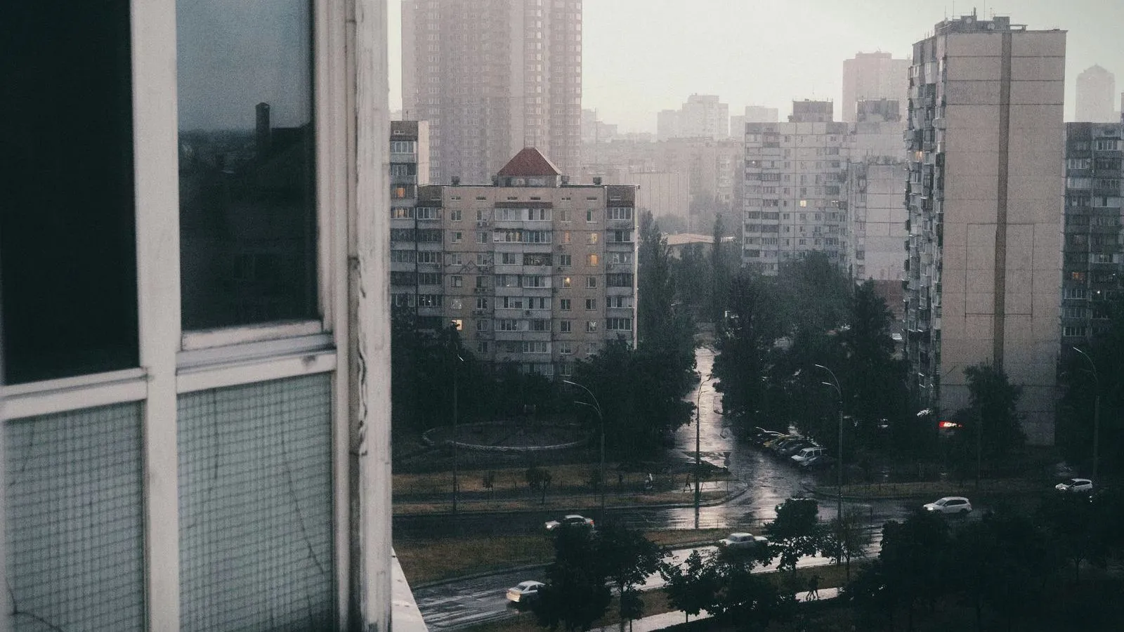 View from an upper window of high-rise apartment blocks and a rain-wet street with cars below on a grey day.