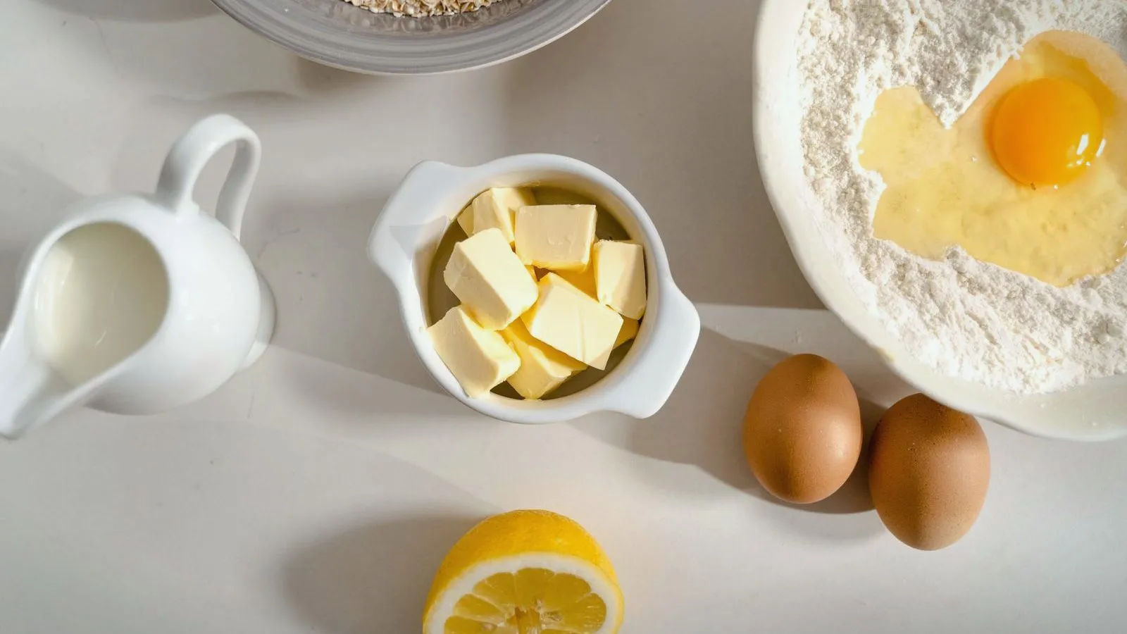 A jug of milk, a bowl of butter cubes, two brown eggs, half a lemon and a bowl of flour with a cracked egg, seen from above on a white counter.