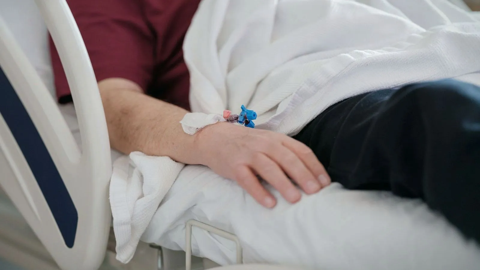 A hand with an intravenous line taped to the back of it rests on white bedding beside a hospital bed rail.