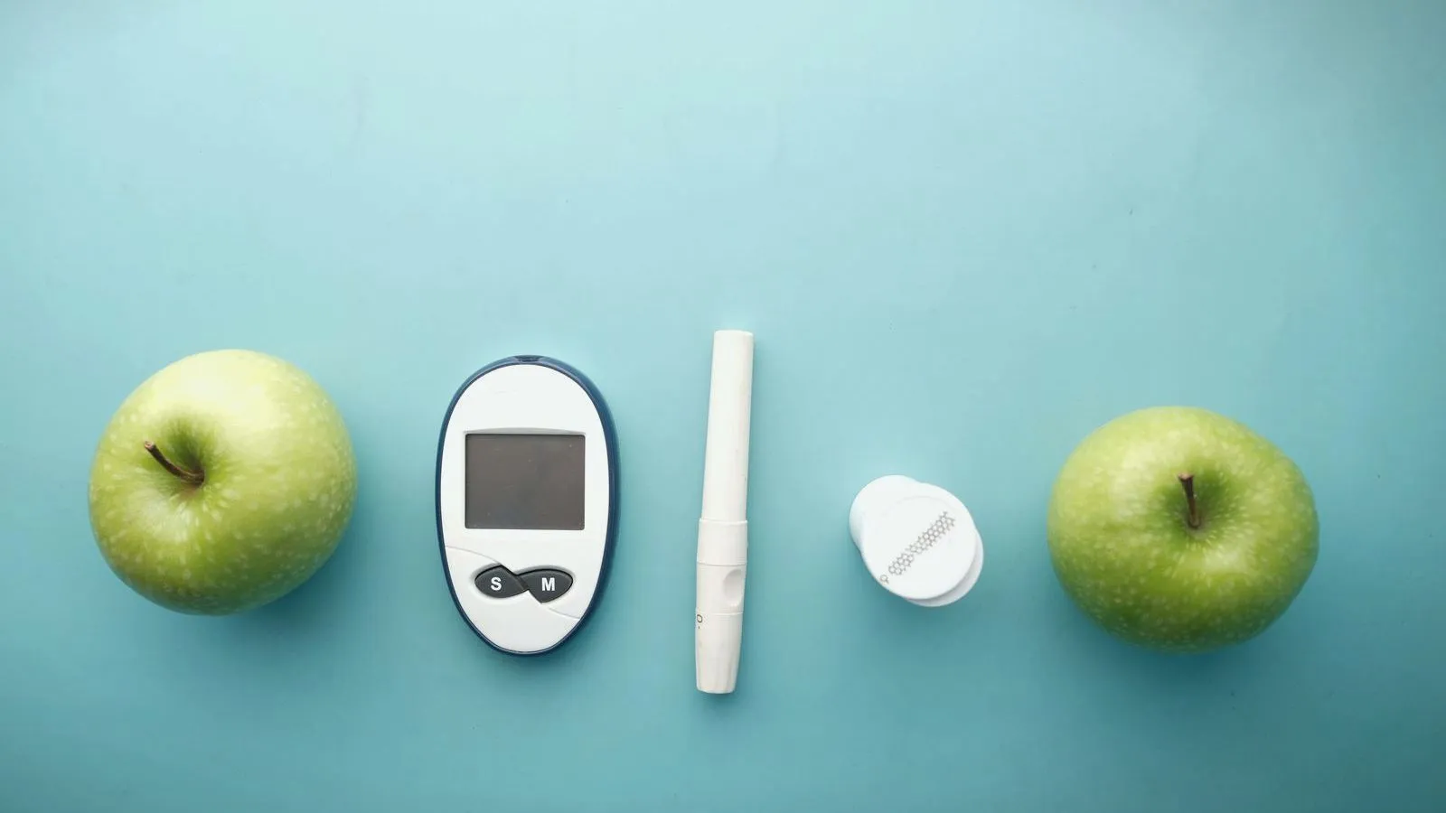 A blood glucose meter, a lancing pen and a test strip container laid in a row between two green apples on a teal background.