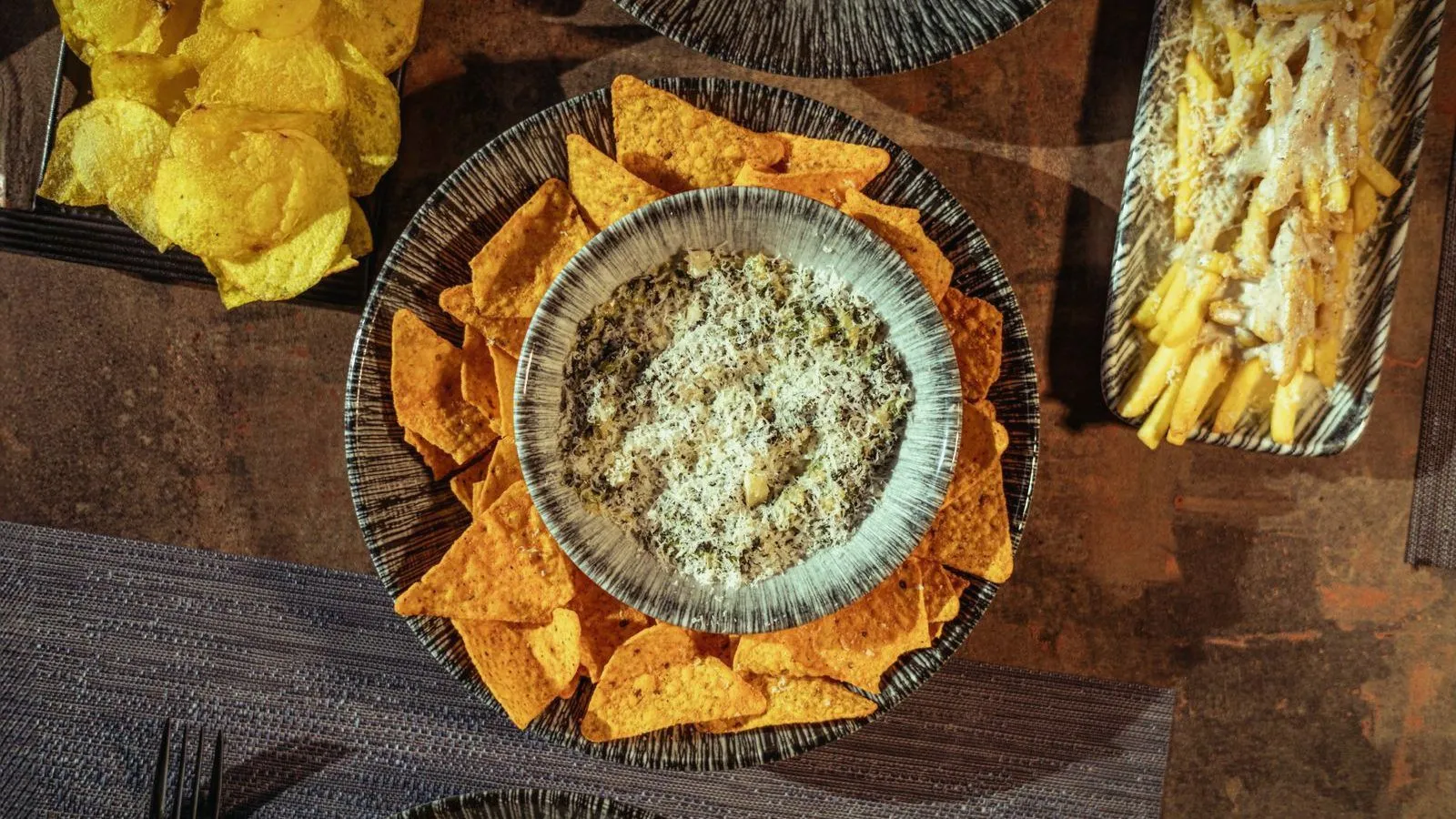 Potato chips, tortilla chips around a bowl of dip and a tray of fries on a table, seen from above.