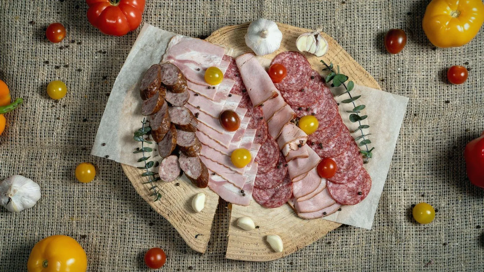 Overhead view of a wooden board of sliced sausage, bacon, ham and salami with cherry tomatoes and garlic on burlap.