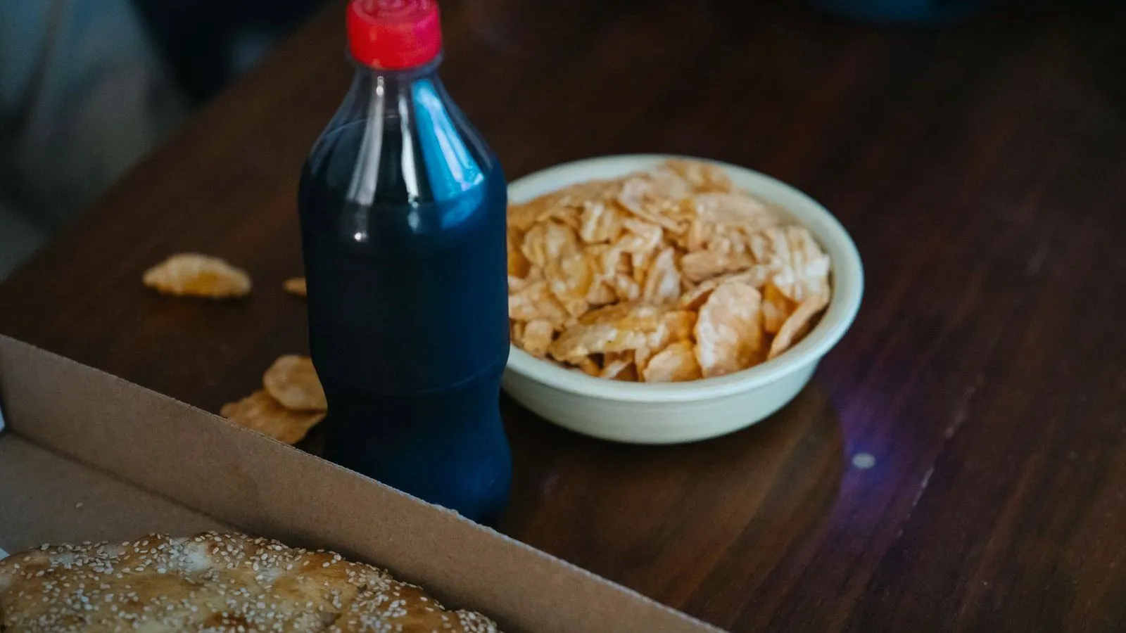 A bottle of dark soda with no label, beside a bowl of potato chips on a wooden table