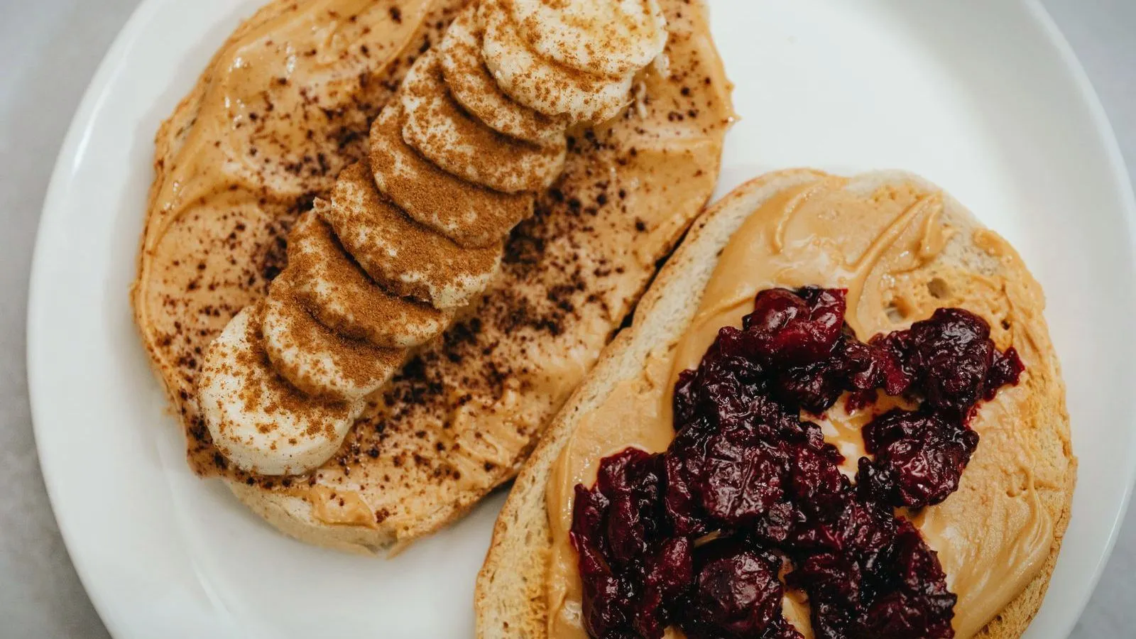 Two slices of toast spread with peanut butter on a white plate, one topped with sliced banana and one with dark berry jam.