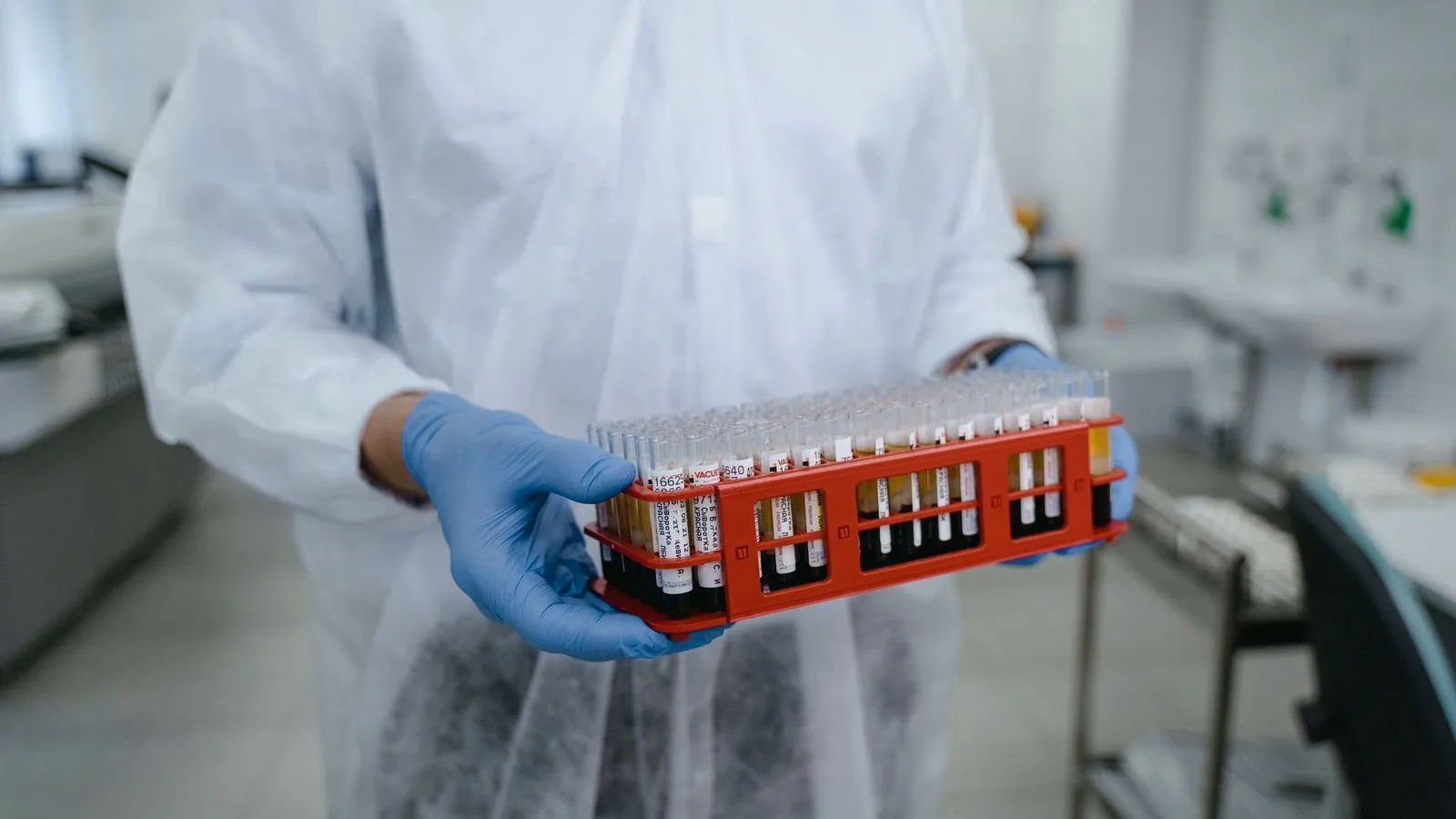 A laboratory worker in a white gown and blue gloves carries a red rack filled with blood sample tubes.
