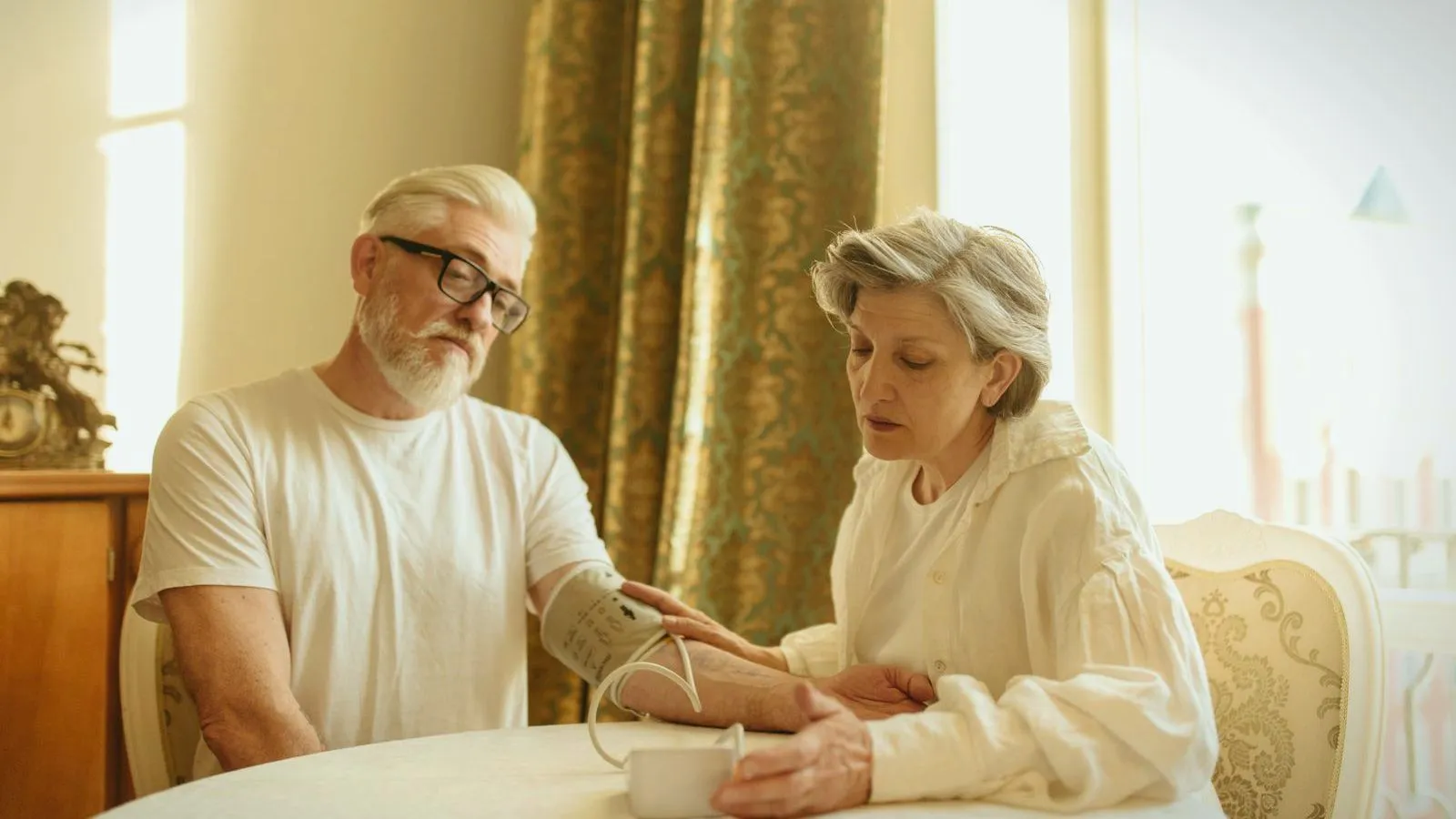 An older man in a white T-shirt sits at a table with a blood pressure cuff on his upper arm while an older woman beside him reads the monitor.