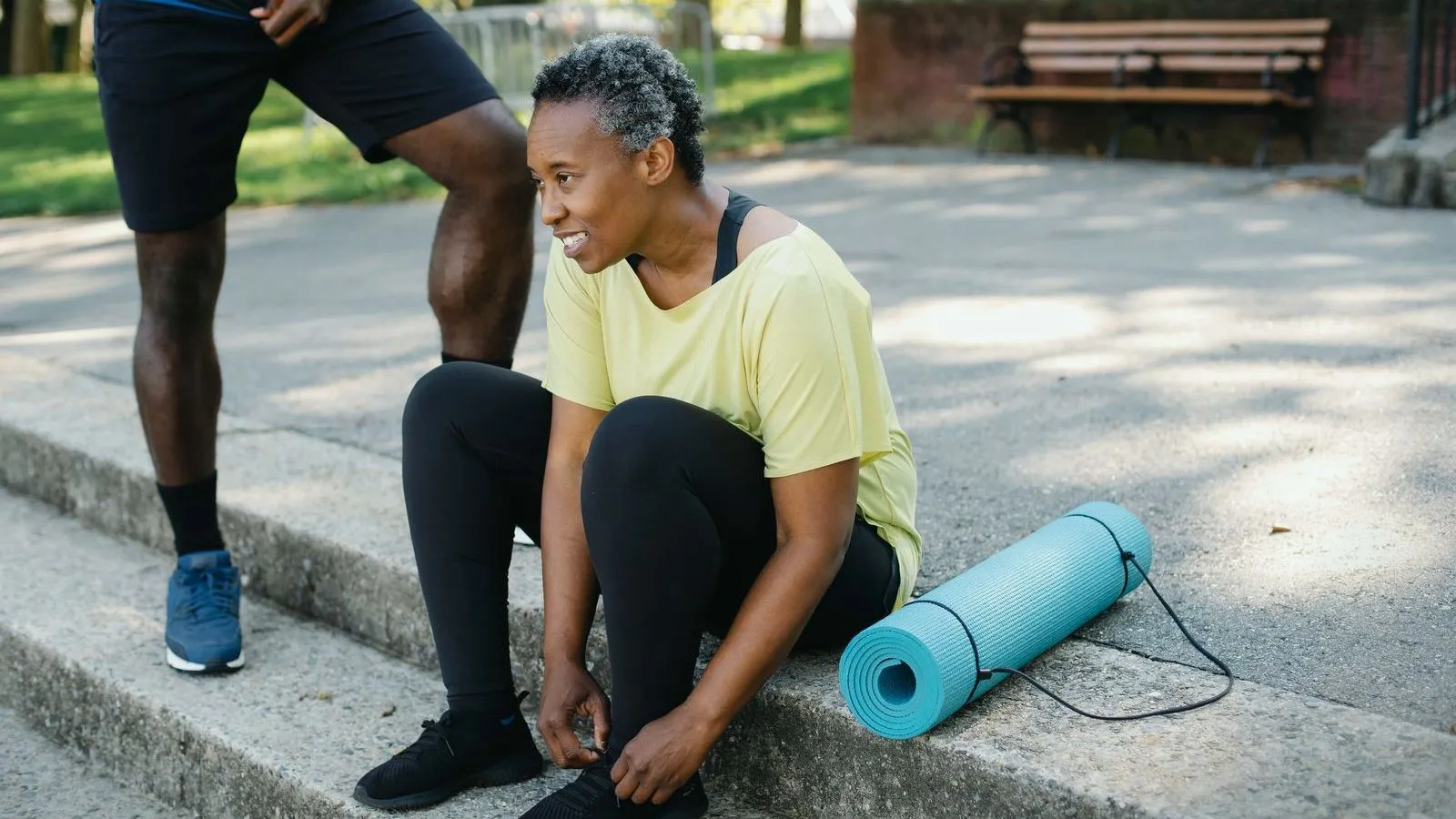 A woman in workout clothes ties a shoelace on park steps beside a rolled yoga mat