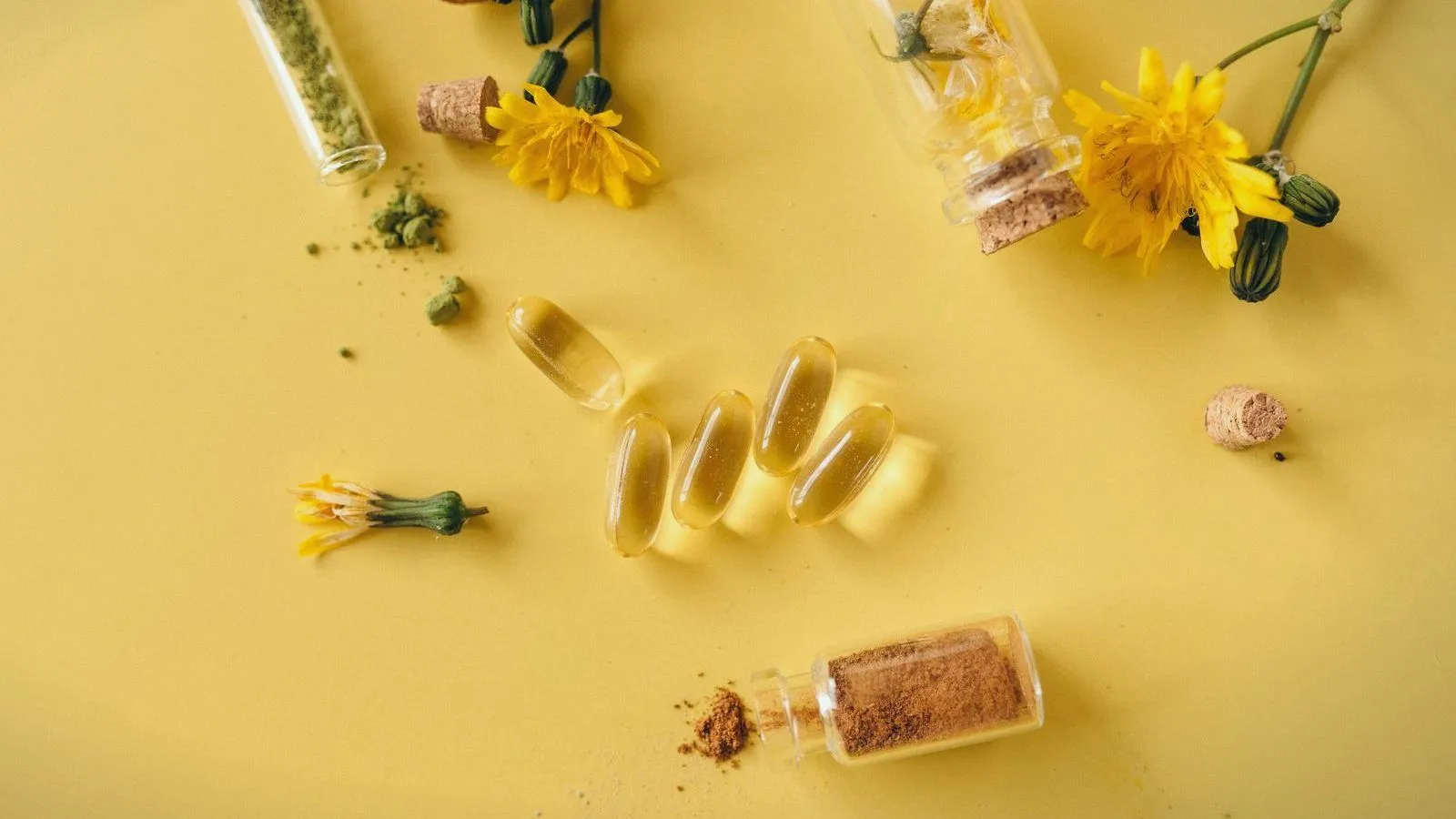 Clear yellow softgel capsules, small corked glass bottles of powder and yellow flowers scattered on a yellow surface.