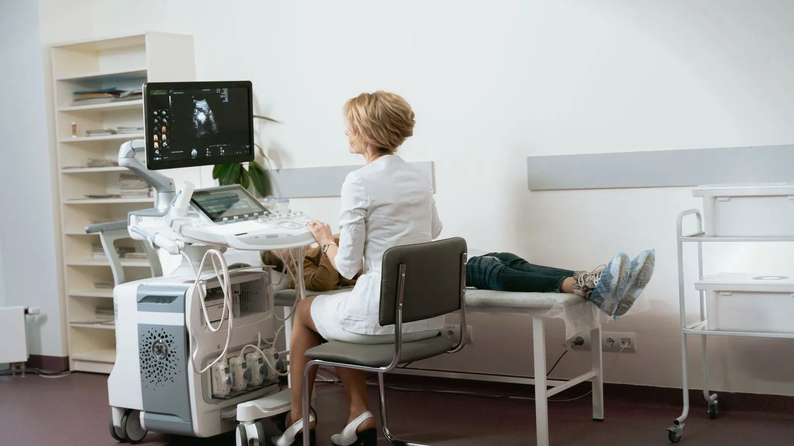 A clinician in a white coat sits at an ultrasound machine beside an exam couch on which a patient is lying, with only the legs in view.