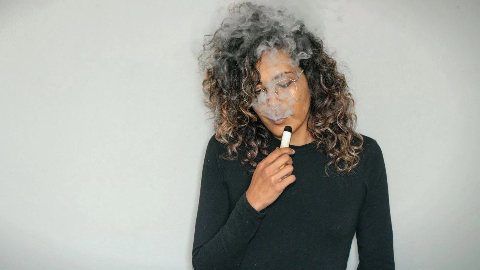 A woman with curly hair, wearing a black top, holds a small white vape to her mouth as vapor drifts around her face.