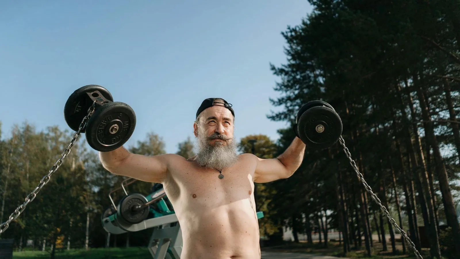 An older man with a gray beard raising two dumbbells outdoors, with pine trees behind.