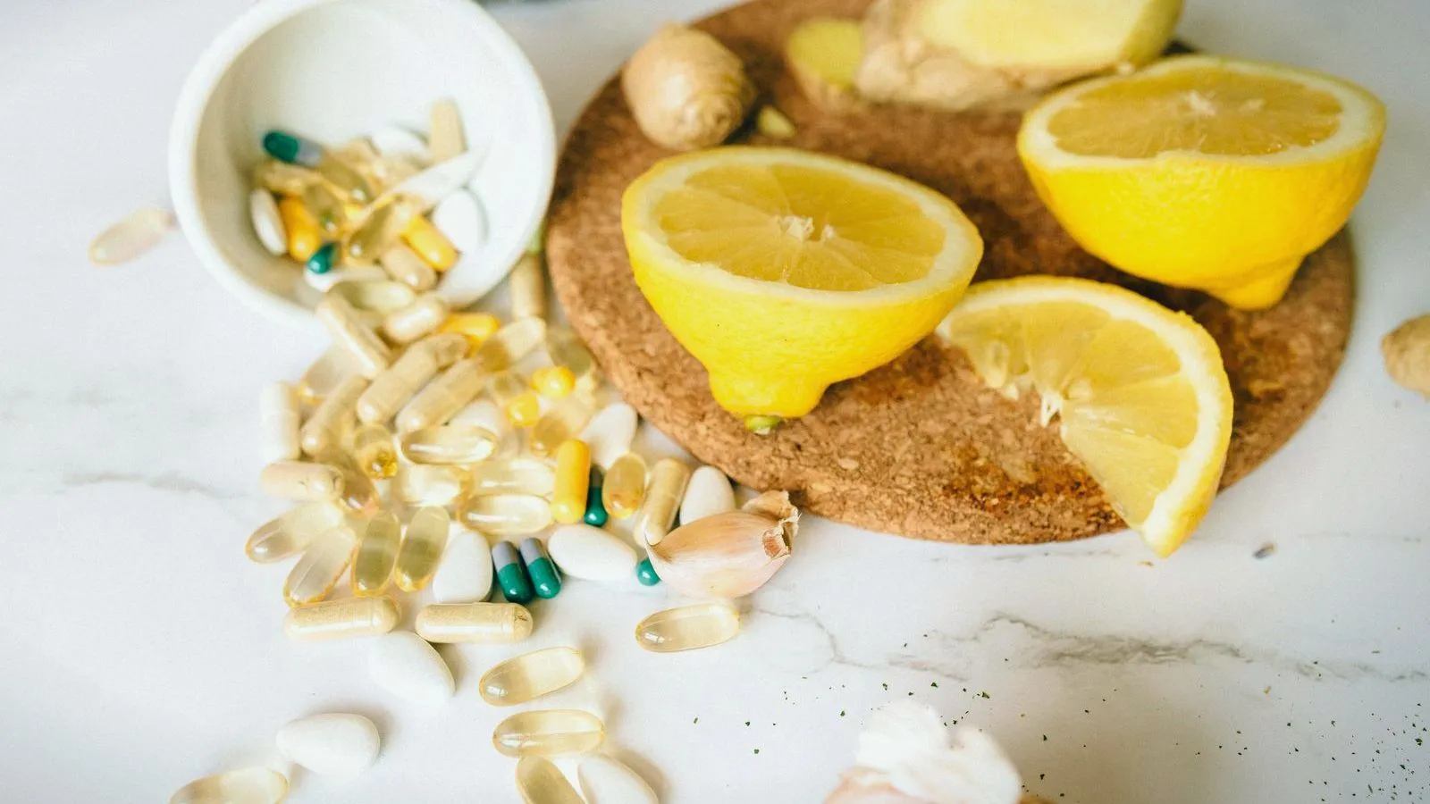 Capsules spilling from a small white bowl beside halved lemons and pieces of ginger on a round cork mat.