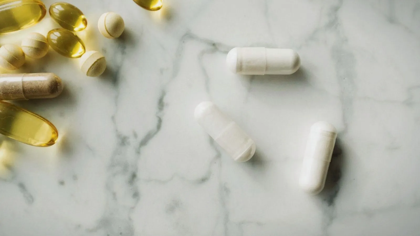White capsules and golden softgels scattered on a marble surface.