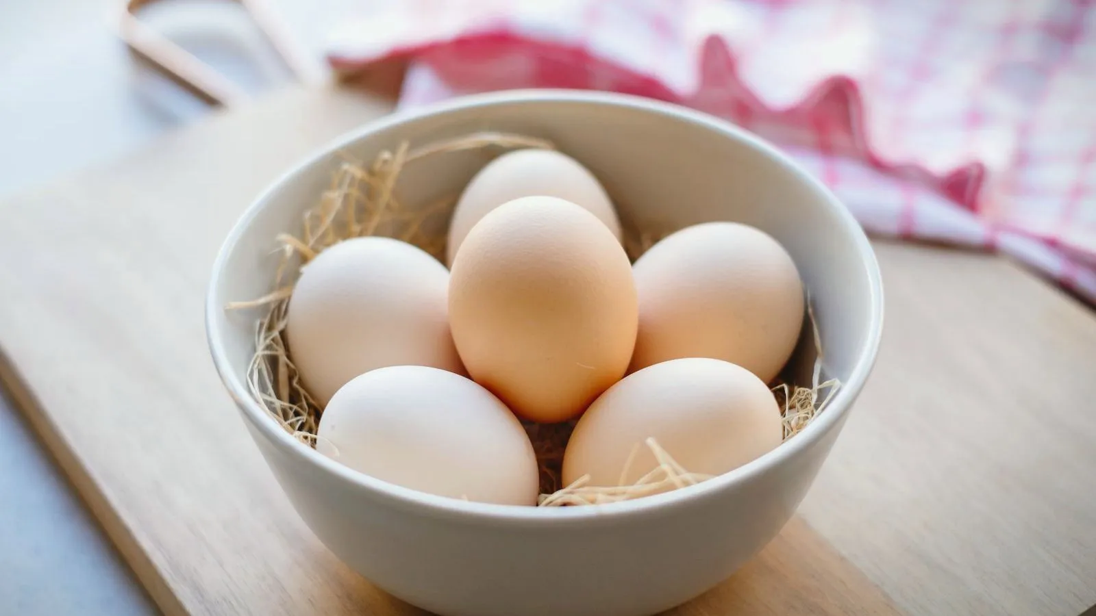 A white bowl of eggs nestled in straw on a wooden board
