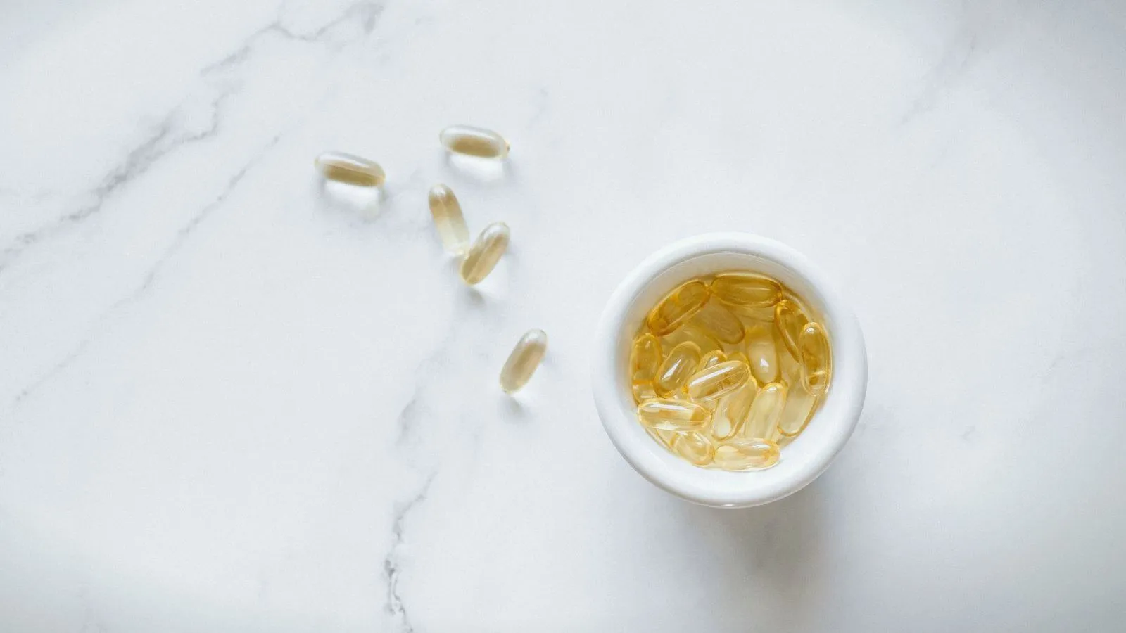 Golden softgel capsules in a small white bowl on marble, a few scattered beside it