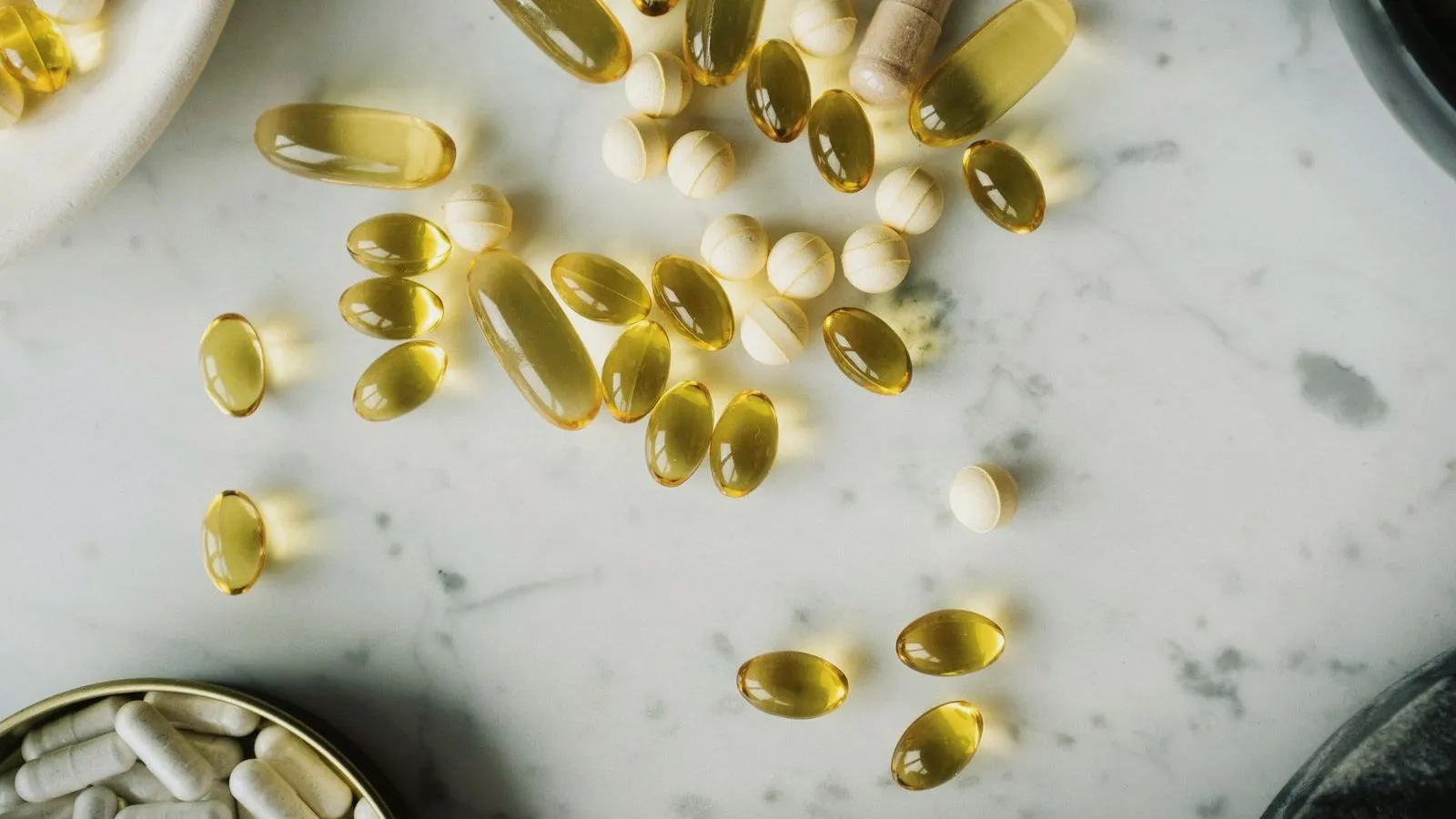 Amber softgel capsules and pale round tablets scattered across a marble surface, with a tin of white capsules at the edge.