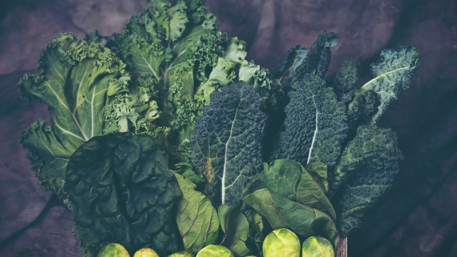 A spread of dark leafy greens including kale and cabbage on a dark surface