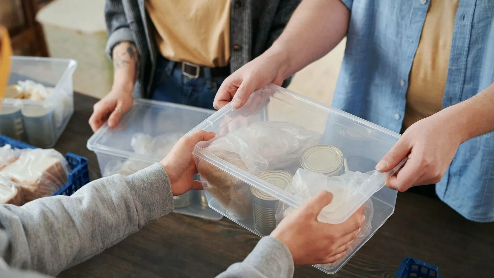 Several pairs of hands passing a clear plastic box of canned food across a table.