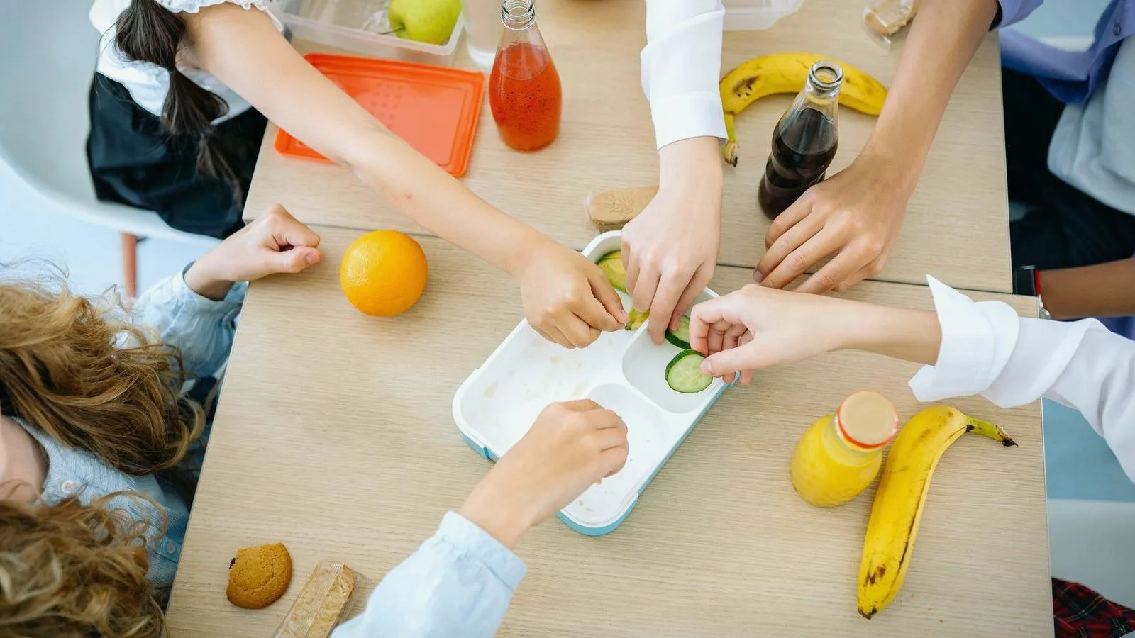 Overhead view of children's hands around a table with cucumber slices, fruit, bananas and bottled drinks.