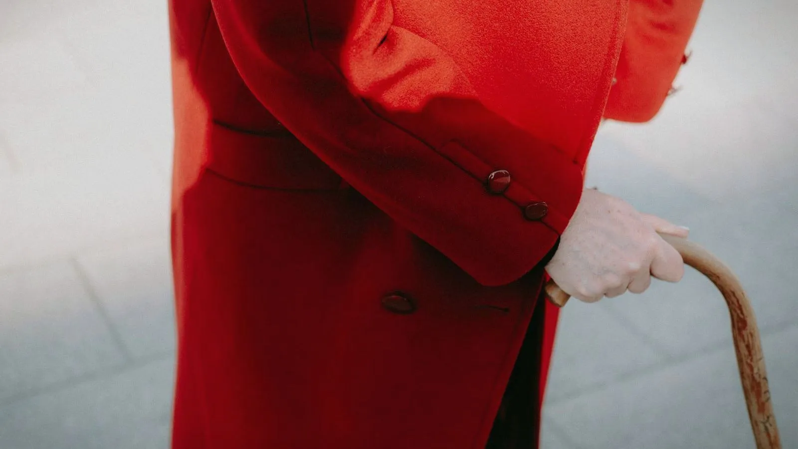 A hand resting on a wooden cane, held by a person in a long red coat whose face is not shown.