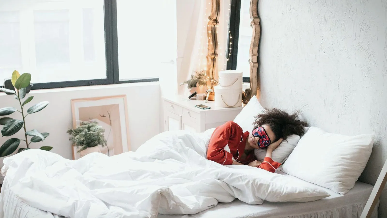A person in red pajamas and a patterned sleep mask asleep in a bed with white covers in a bright, sunlit room.