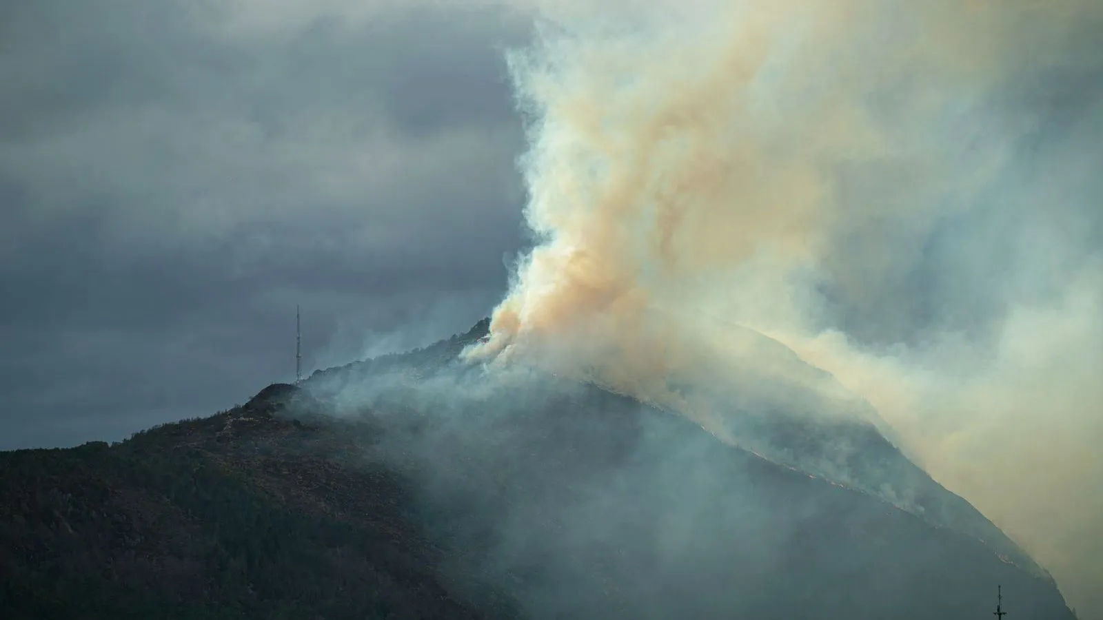 Thick smoke rises from a forested mountainside and spreads across a dark sky.