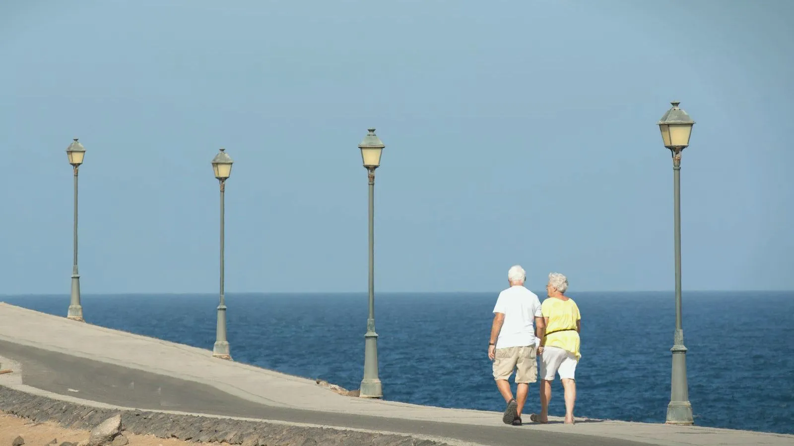 An older couple in shorts, seen from behind, walking along a seaside promenade lined with lamp posts.