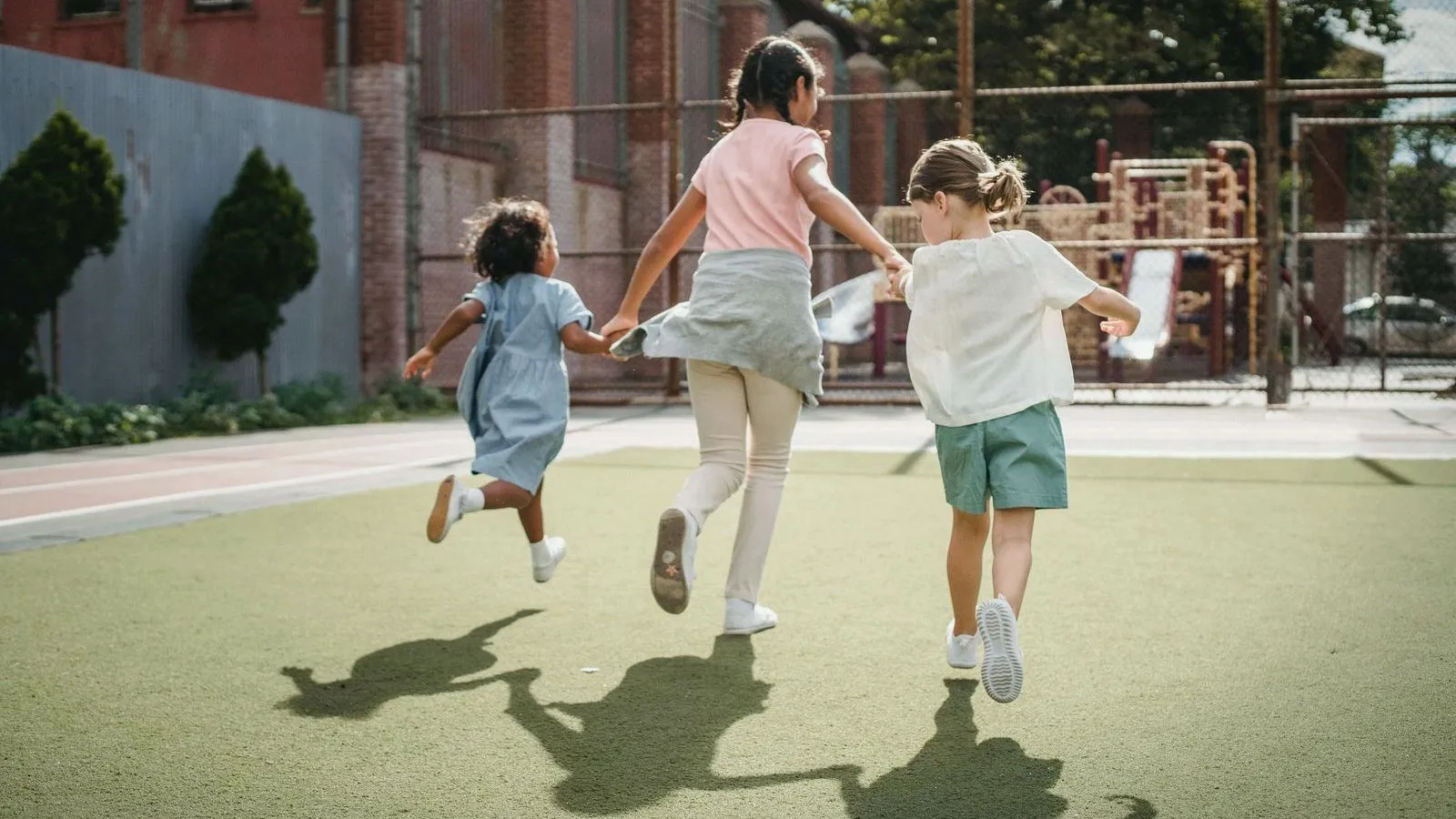 Three children seen from behind run hand in hand across a schoolyard toward a playground, casting long shadows.