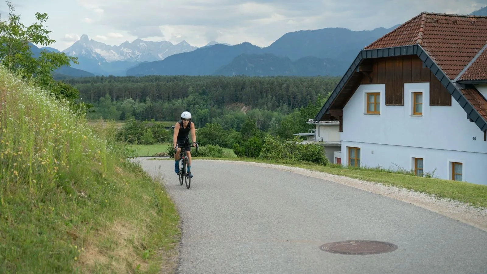 A cyclist riding along a quiet road past a white house, with forest and mountains behind.